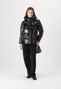 Chaqueta puffer negra y brillante con capucha, que presenta un cierre de cremallera y un diseño acolchado; combinada con pantalones negros y un bolso pequeño.