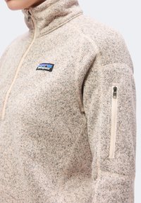 Μπεζ πλεκτό fleece Patagonia με φερμουάρ τεταρτημόριου και φερμουάρ τσέπης στο αριστερό πάνω μανίκι, που φοριέται από έναν άνθρωπο.