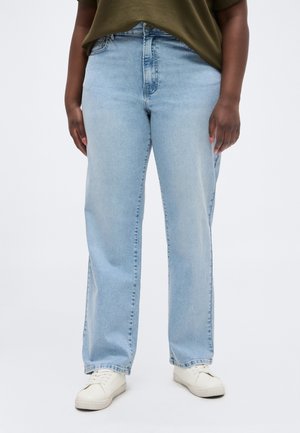 Személy világoskék, egyenes szárú farmernadrágot, olívazöld felsőt és fehér tornacipőt visel, fehér háttér előtt áll.