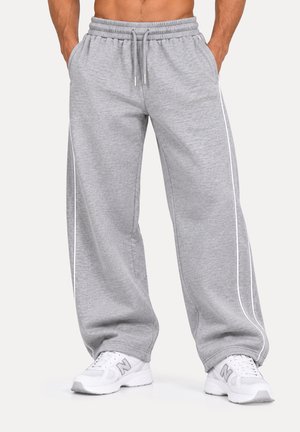 Modelo con pantalones de chándal grises holgados con cordón y ribetes blancos en los laterales, y zapatillas blancas, con las manos en los bolsillos.