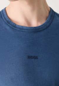 Gros plan d'une personne portant un t-shirt bleu à col rond avec le mot BOSS brodé en bleu foncé sur la poitrine.