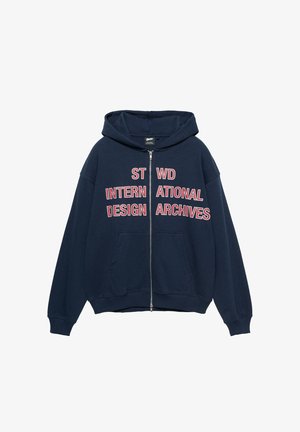 Marineblauer Kapuzenpullover mit Reißverschluss, Fronttaschen und rot-weißem Blocktext mit der Aufschrift "ST WD INTERNATIONAL DESIGN ARCHIVES" auf der Brust.
