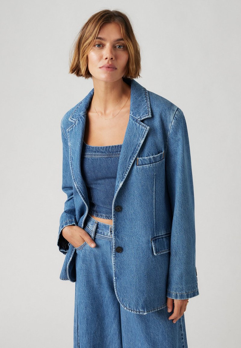 Denim blazer v svetlo modri barvi z izrezanim reverjem, enakim gumbom za zapiranje in žepom na obliž. Ujemajoč denim top.