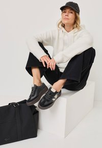 Sneaker nera e grigia con una tomaia testurizzata e suola in gomma. Il design presenta lacci e un accento chiaro a contrasto. Una borsa nera è nelle vicinanze.