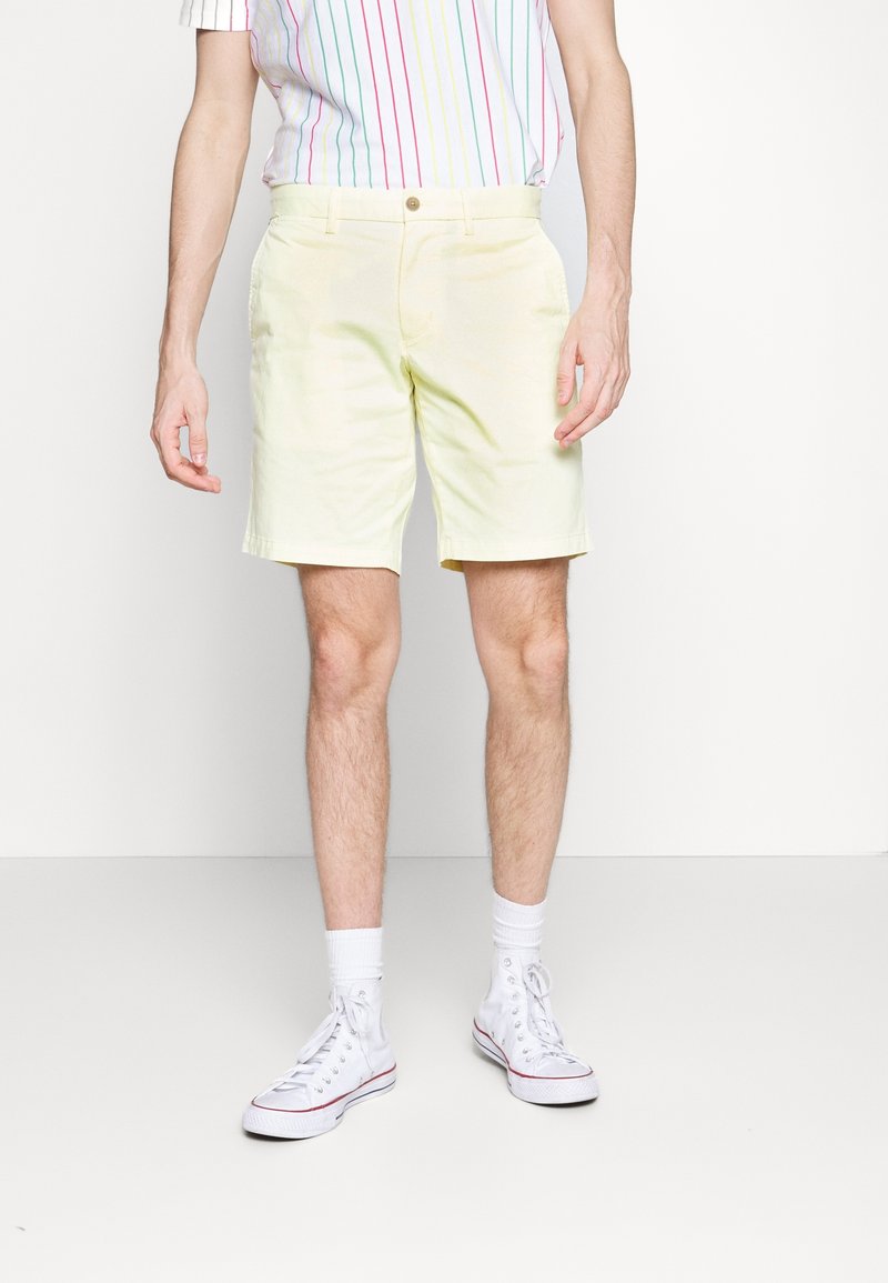 Homme portant un short jaune clair, des baskets montantes blanches, des chaussettes blanches et une chemise blanche à rayures verticales multicolores, debout devant un fond uni.