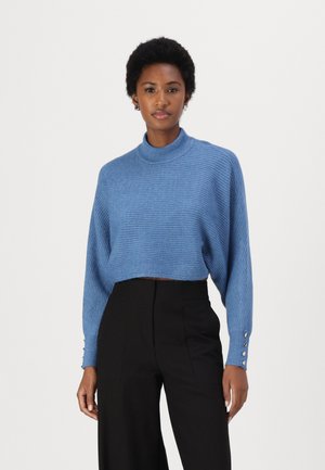 Blue ribbed cropped sweater with a high collar and long sleeves, featuring button accents on the cuffs. Paired with black high-waisted trousers.