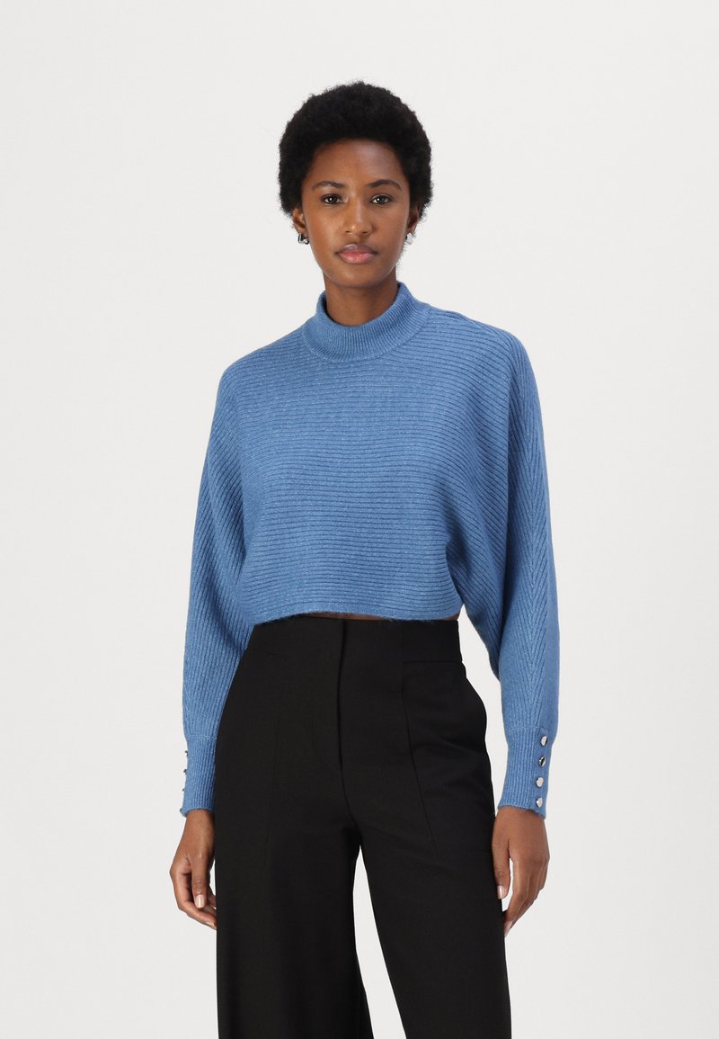 Maglione corto azzurro a coste con colletto alto e maniche lunghe, dotato di bottoni decorativi sui polsini. Abbinato a pantaloni neri a vita alta.