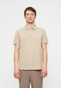 Bărbat stând cu fața înainte, purtând un tricou polo bej cu mâneci scurte și pantaloni maro pe un fundal alb simplu.