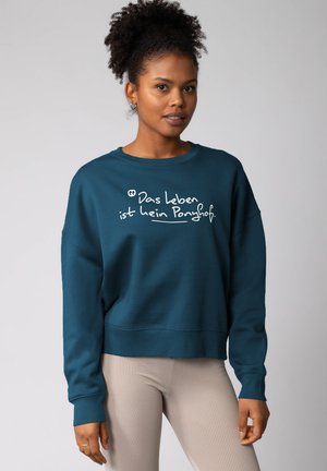 Teal cropped sweatshirt met lange mouwen, voorzien van witte tekst: "Das Leben ist kein Ponyhof." Gekocht met geribbelde beige legging.