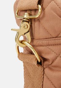 Broche de metal dorado unido a una correa de tela color camel y esquina acolchada de un bolso con costuras visibles y detalle de cremallera.