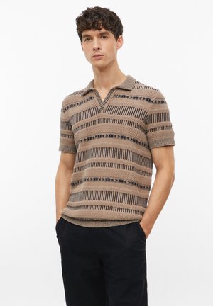 Jonge man met krullend haar draagt een beige en zwart gestreept gebreid poloshirt met korte mouwen en een zwarte broek, handen in de zakken.