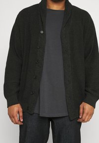 Dunkelgrüne Strickjacke mit Schalkragen, ausgestattet mit sechs schwarzen Knöpfen, getragen über einem grauen Hemd. Strukturiertes Gewebe mit geripptem Design.