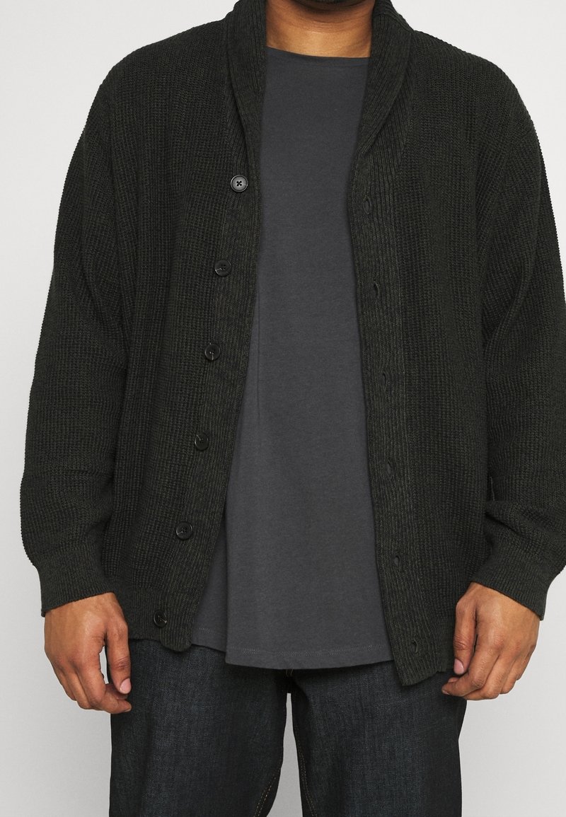 Dunkelgrüne Strickjacke mit Schalkragen, ausgestattet mit sechs schwarzen Knöpfen, getragen über einem grauen Hemd. Strukturiertes Gewebe mit geripptem Design.