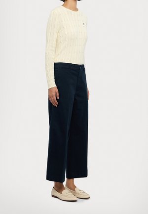 Roomkleurige kabelgebreide trui, wijde marineblauwe broek en beige loafers. De outfit heeft een textuurrijke stof en een ontspannen silhouet. Kenmerkende geribde patronen.