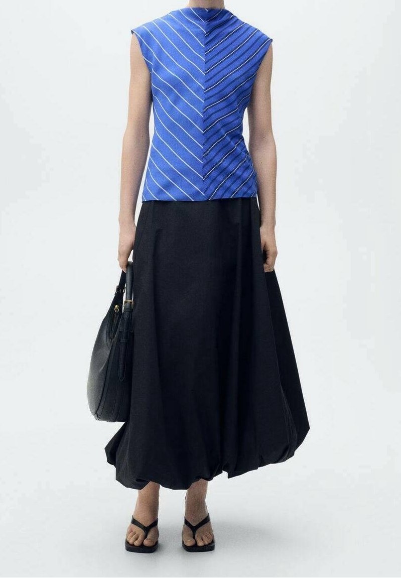 Femme portant un haut rayé bleu sans manches et une longue jupe noire fluide, tenant un sac à main noir et portant des sandales noires.