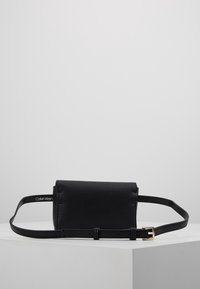 Černá kožená pásková taška s texturovaným povrchem, vybavená dlouhým nastavitelným popruhem a sponou v zlatém tónu. Viditelné logo značky Calvin Klein.