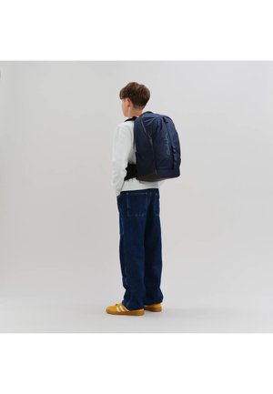 Junge trägt ein weißes Sweatshirt, blaue Jeans, gelbe Turnschuhe und einen dunkelblauen Rucksack und steht vor einem einfarbigen hellgrauen Hintergrund.