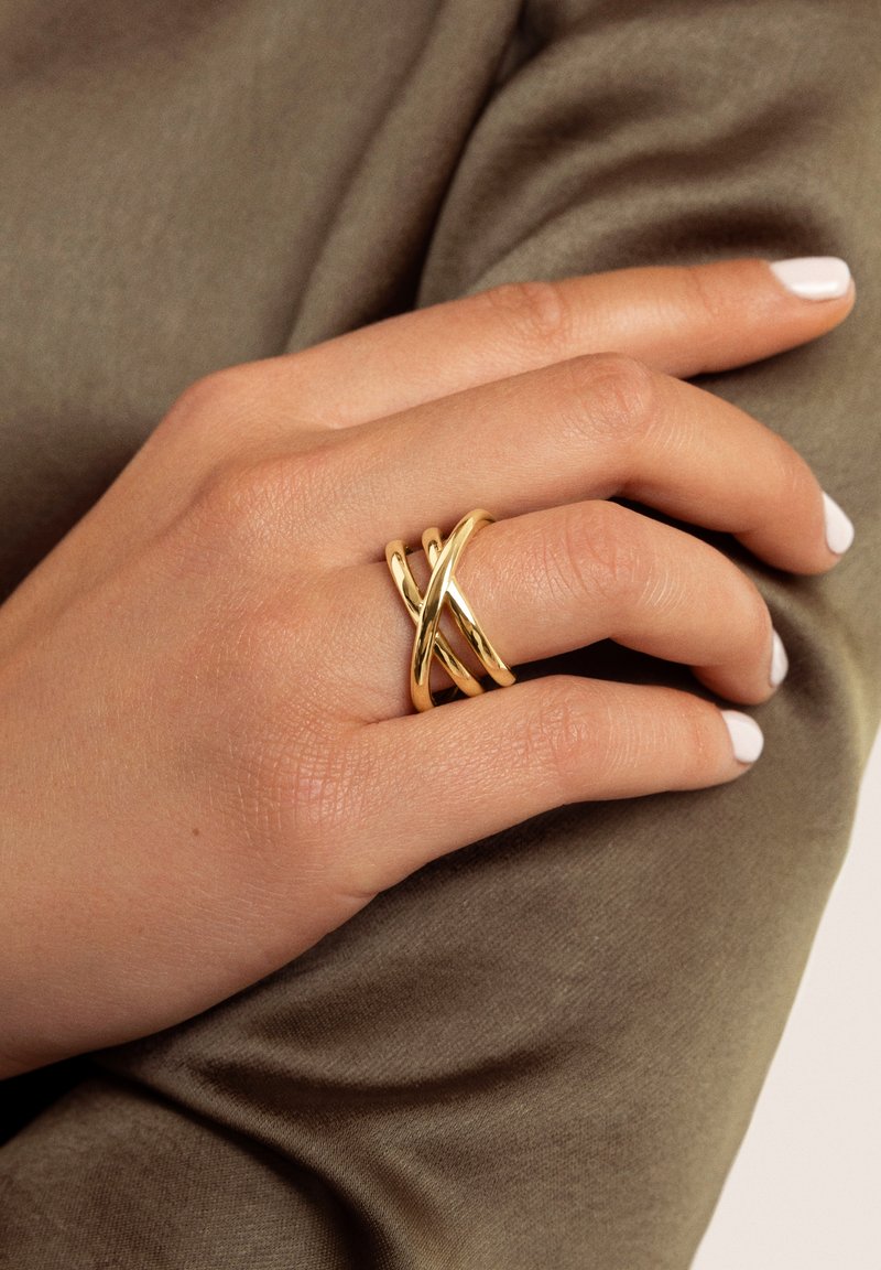 Anillo de metal dorado con un diseño entrelazado y superficies lisas y brillantes. La mano descansa sobre un fondo texturizado de tela verde oliva.