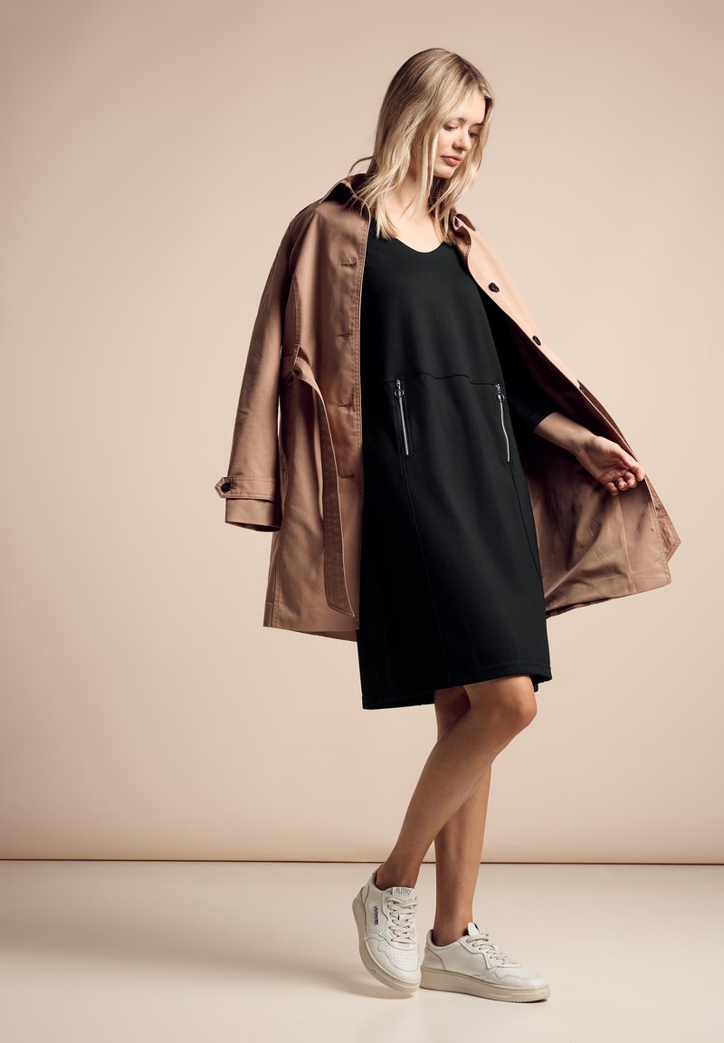 Vestito nero con tasche anteriori con zip, indossato sotto un trench rosa chiaro. Sneakers bianche completano l'outfit. Tessuti morbidi, vestibilità rilassata.