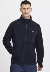 Svart fleece-jakke med høy krage, glidelåslukking, brystlomme og sidelommer. Har lange ermer og en myk tekstur.