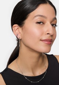 Boucles d'oreilles en argent avec des détails pendants et un collier en chaîne argenté assorti orné de petits accents. Le modèle porte un haut noir.