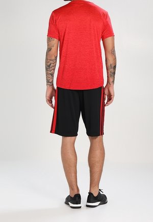 Hombre con camiseta roja, pantalones cortos negros con rayas rojas y zapatillas negras, de pie con la espalda hacia la cámara sobre un fondo liso.