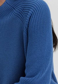 Stickad tröja i mörkblått, med en texturerad mönster, ribbad halsringning och långa ärmar. Materialet verkar mjukt och stretchigt.