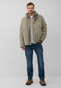 Man in een beige gewatteerde jas over een geruit shirt en een witte t-shirt, blauwe jeans en bruine veterschoenen, staand tegen een effen achtergrond.