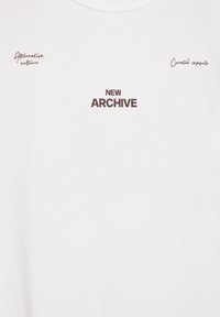 Valkoinen puuvillapaita, jossa on ruskea teksti: "UUSI ARKISTO", "Kuraattori kapseli" ja "Vaihtoehtokulttuuri". Pyöreä kaula-aukko, yksinkertainen muotoilu.