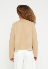 Kobieta o kręconych włosach, ubrana w beżową zamszową kurtkę i białe spodnie, pokazana od tyłu na jednolitym białym tle.