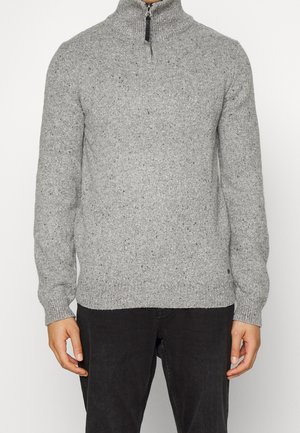 Mężczyzna ubrany w szary melanżowy sweter z dzianiny z półgolfem na zamek i czarne spodnie, stojący na białym tle.