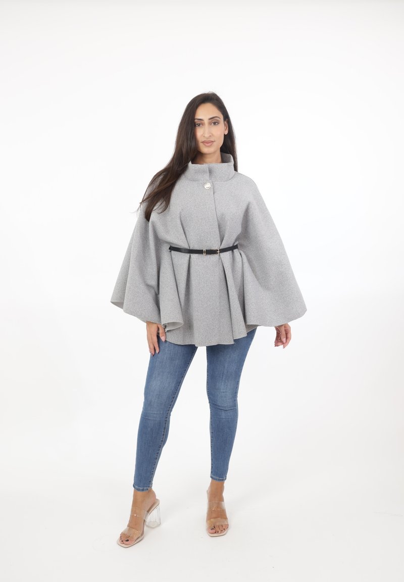 Capa de lana gris con un cuello alto, cierre frontal de botones y un cinturón negro en la cintura, superpuesta sobre jeans ajustados azules y tacones transparentes.