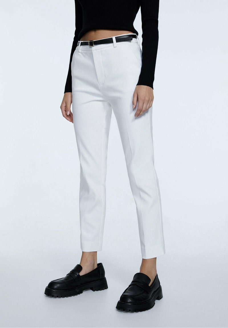 Pantalones blancos ajustados con un corte slim, con trabillas para cinturón y un cinturón de cuero negro. Combinados con mocasines negros gruesos. Textura suave, sin patrones.