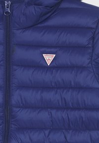 Marineblå polstret vest med lynlås og horisontal quiltet tekstur. Har et trekantet rødt og hvidt "Guess" logo.