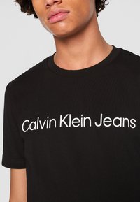Osoba nosząca czarną koszulkę z białym tekstem "Calvin Klein Jeans" na piersi, pokazana od szyi do połowy tułowia.