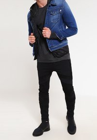 Mann trägt ein dunkelgraues Hemd, blaue Jeansjacke, schwarze schmale Hose und schwarze Slipper vor einem einfarbigen hellen Hintergrund.