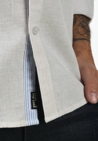 Hvid linnedskjorte med lyseblå stribet inderfor. Har en knap ved kraven og et mærke, der står "WEAR THE CODE." Nærbillede.