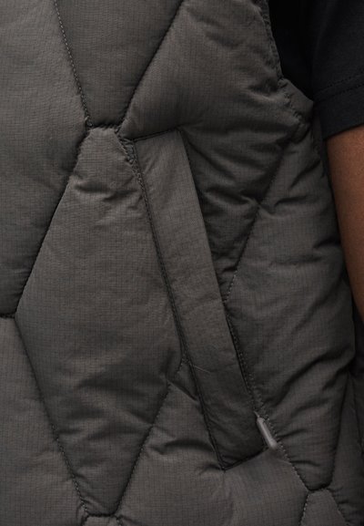 Colete cinzento acolchoado feito de tecido texturizado com padrão geométrico de diamantes, com bolso lateral e ferragens visíveis mínimas.