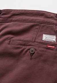 Bordo tkanine hlače s glatkom teksturom, s stražnjim džepom, kopčanjem na gumb i bijelom etiketom s "Levi's" brendiranjem.