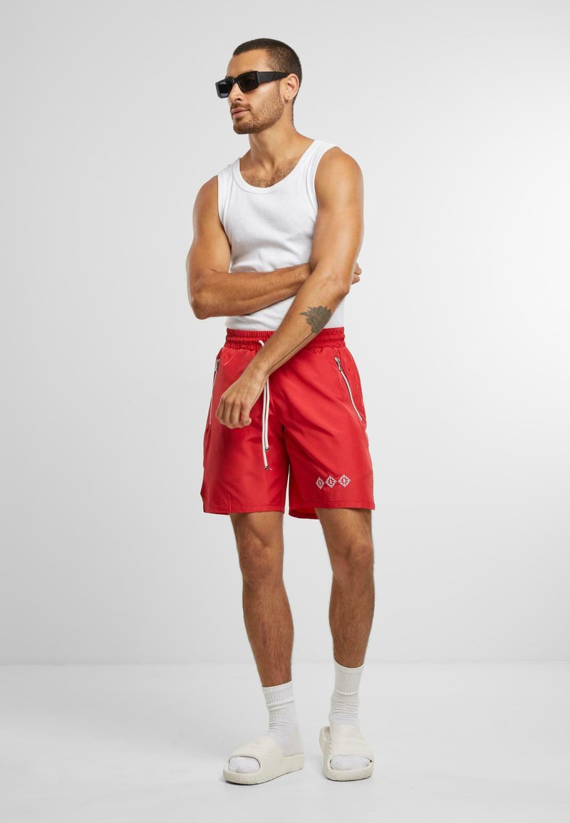 Pantalones cortos rojos con cordón, dos cremalleras laterales y un patrón de diamante bordado en blanco. Combinados con una camiseta sin mangas blanca y sandalias.