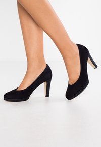 Svarta mockaskor med hög klack och rund tå, med en glittrig svart plattform och hälaccent. Slät textur och klassisk design.