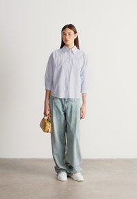 Ανοιχτό μπλε πουκάμισο με γιακά και μακριά μανίκια, συνδυασμένο με χαλαρά τζιν σε τυρκουάζ απόχρωση. Το μοντέλο κρατά μια μικρή κίτρινη τσάντα και φοράει λευκά αθλητικά παπούτσια.