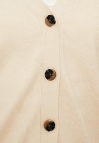 Μπεζ πουλόβερ με υφή υφάσματος, με σχέδιο V-λαιμόκοψης και τέσσερα στρογγυλά μαύρα και καφέ μαρμάρινα κουμπιά κατά μήκος του μπροστινού μέρους.