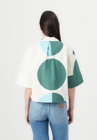 Chemise à manches courtes blanche avec de grands motifs circulaires verts et bleu clair, dotée d'un pli au dos et d'un col classique. Associée à un jean bleu.