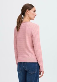 Rosa strikket genser med teksturert kabeldesign, rund hals og lange ermer. Bunnpartiet har ribbestrikket mansjett, kombinert med blå jeans.