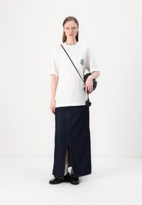 White cotton t-shirt with short sleeves and green graphic print, paired with a long dark denim skirt featuring a front slit, and black shoes.
