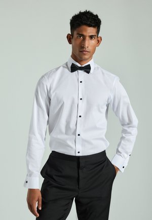 Fiatal férfi fehér ingben, fekete gombokkal és fekete csokornyakkendővel, fekete nadrágot viselve, áll, egyik kezét a zsebében tartva.