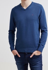 Granatowy sweter z dzianiny z dekoltem w serek, ściągaczami na rękawach i spodzie, z małym logo na piersi. Gładka faktura, dopasowany krój.