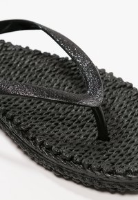 Primer plano de una sandalia tipo chancla negra tejida con tiras brillantes texturizadas y un patrón trenzado en la suela sobre un fondo blanco.