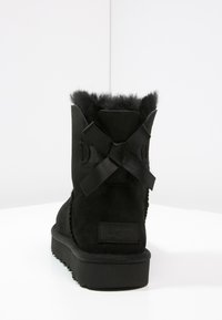 Bottes en suède noir avec un tige courte, dotées d'un doux doublage en fourrure, de nœuds en satin noir décoratifs à l'arrière, et d'une semelle noire texturée.